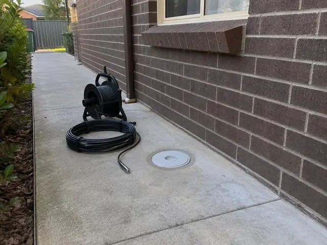 Capped drain inspection opening beside a coiled camera reel on a concrete side path by a brick house in Kellyville