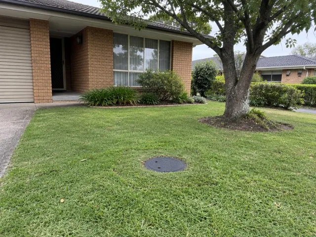 Flush circular sewer cap in a neat front lawn beside a brick house in Kellyville